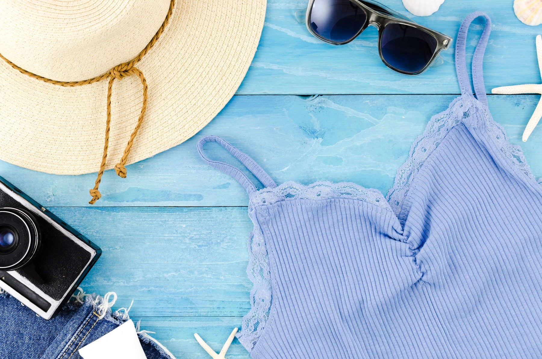 Vintage summer outfit featuring a blue tank top, straw hat, sunglasses, and a retro camera on a blue wooden background.