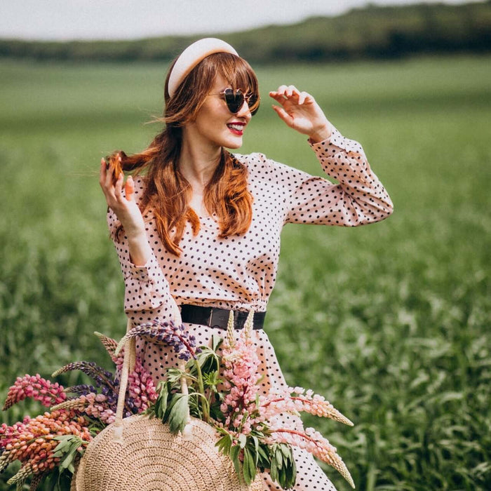 Woman in polka dot dress wearing sunglasses and headband, smiling and holding flowers in a field of green grass during spring/summer season