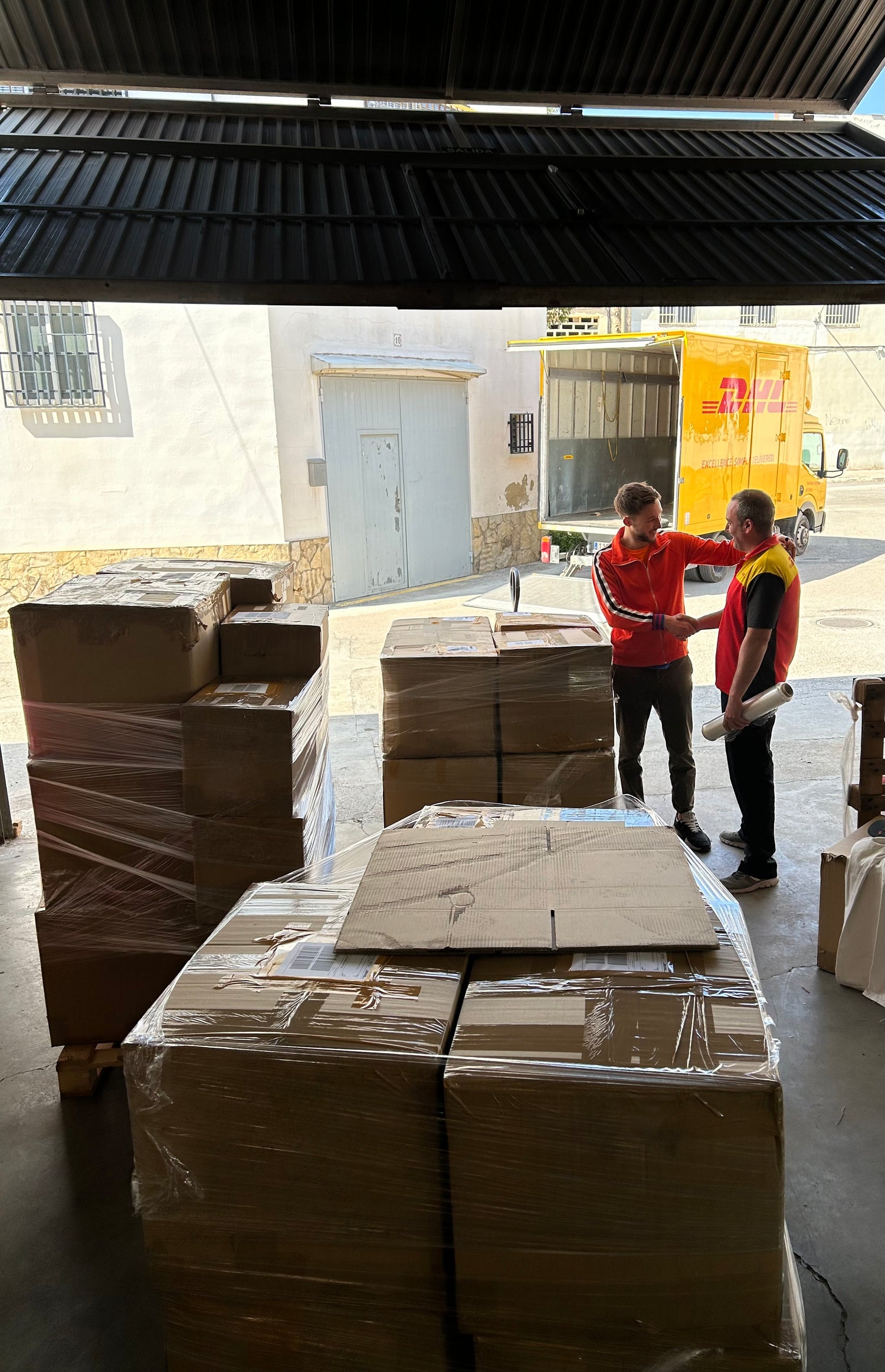 Workers discussing logistics next to stacked cardboard boxes in a warehouse with a delivery truck in the background.