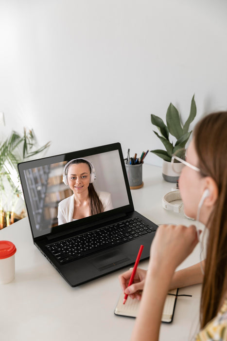 Woman attending a virtual video meeting and taking notes while looking at laptop screen with another woman wearing headphones.