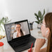 Woman attending a virtual video meeting and taking notes while looking at laptop screen with another woman wearing headphones.